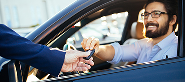 Man in car smiling while a man outside of the car is handing in a key to the vehicle through the drivers window. 